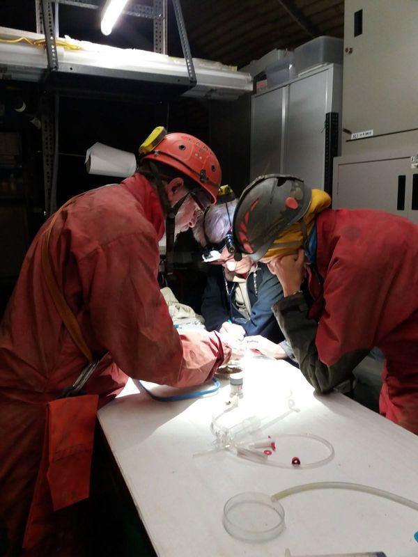 Tre ricercatori in tuta rossa e casco esaminano campioni su un banco illuminato